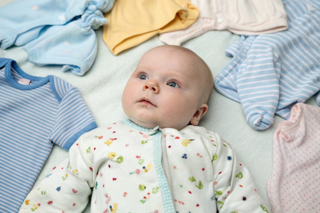 Baby lying on a soft blanket surrounded by colorful baby clothes