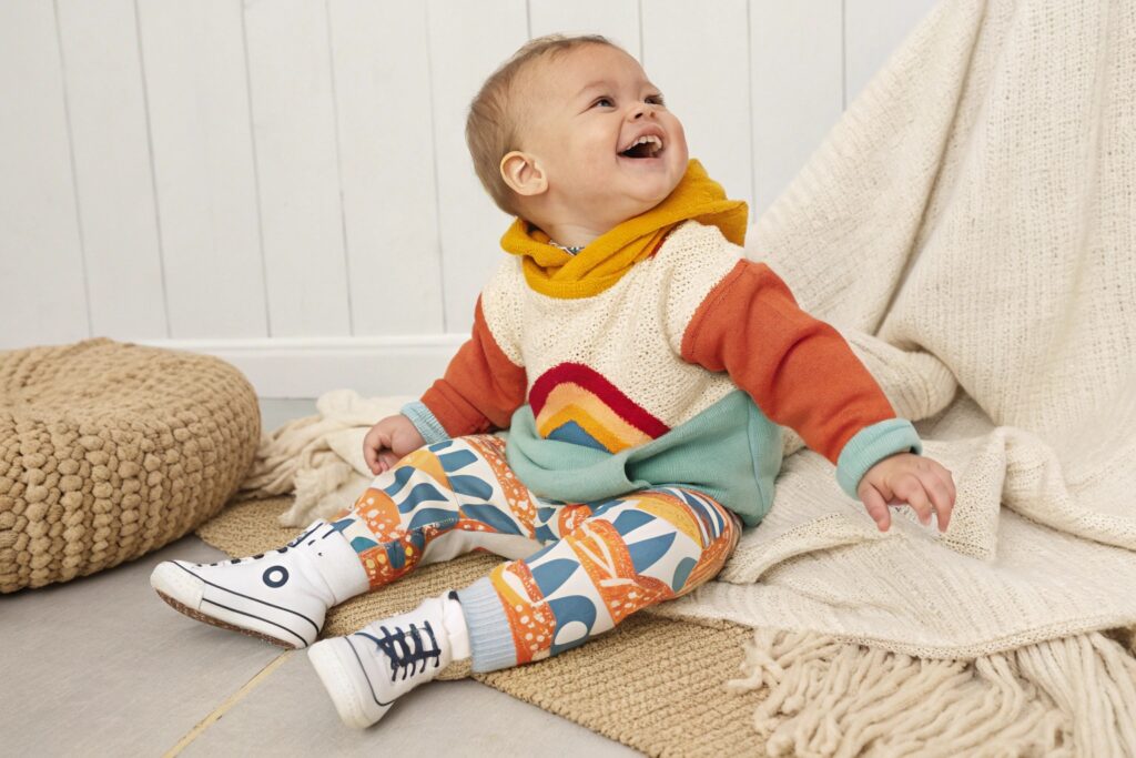 Happy baby wearing a colorful hoodie and patterned pants, sitting on a cozy mat